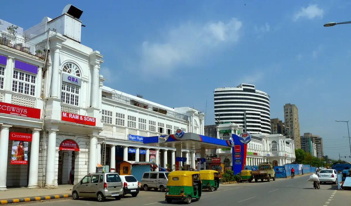 How can I reach Jaipur? 3 Colonial-style white buildings of Connaught Place in New Delhi, lined with shops, vehicles, and a fuel station under a clear blue sky.