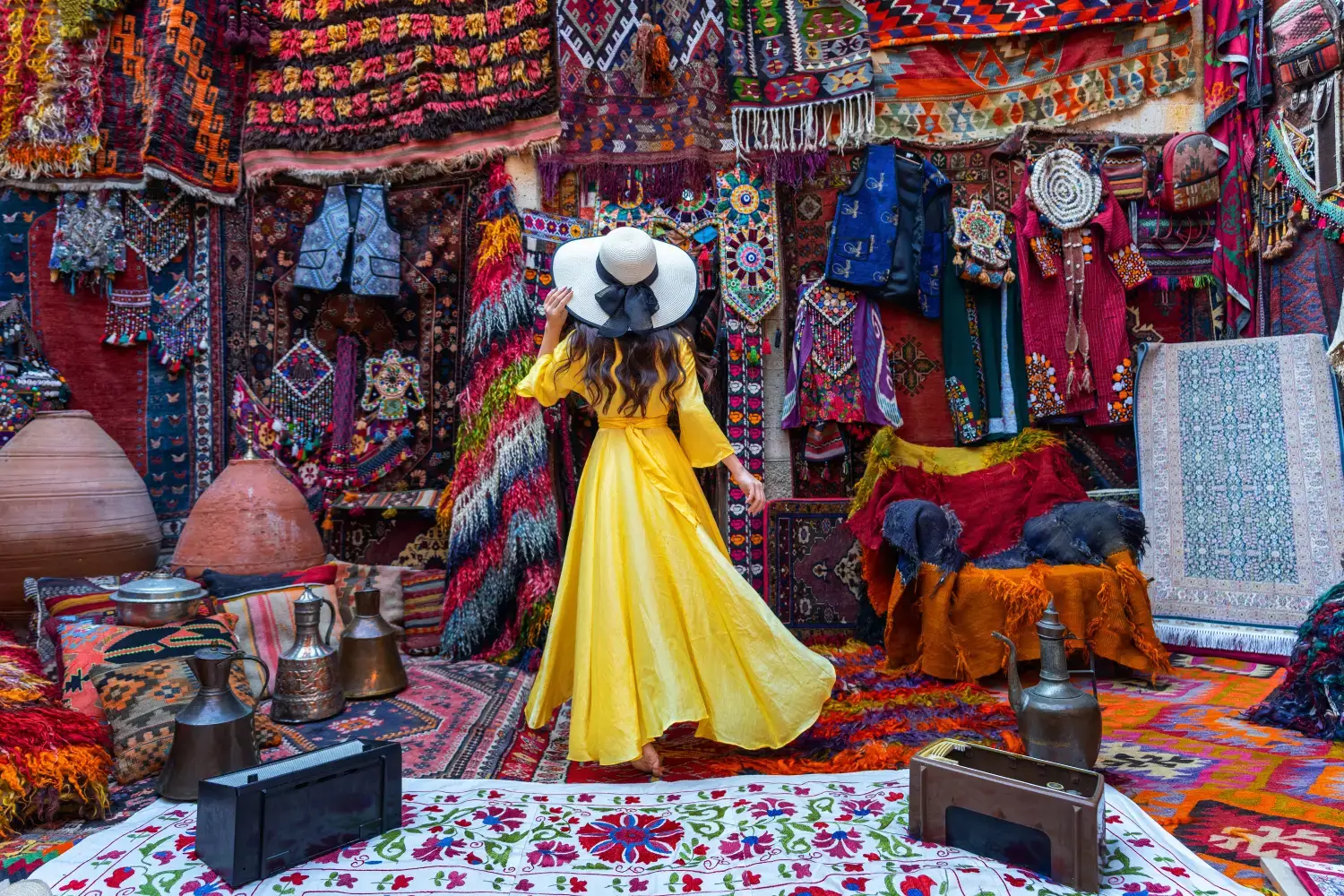 What unique experiences should I try in Jaipur? 1 A guest wearing a vibrant yellow dress and a wide-brimmed hat explores a colorful market filled with traditional Indian rugs, textiles, and artifacts during their India Group Tour and Golden Triangle Tour provided by Travel with Vasco.