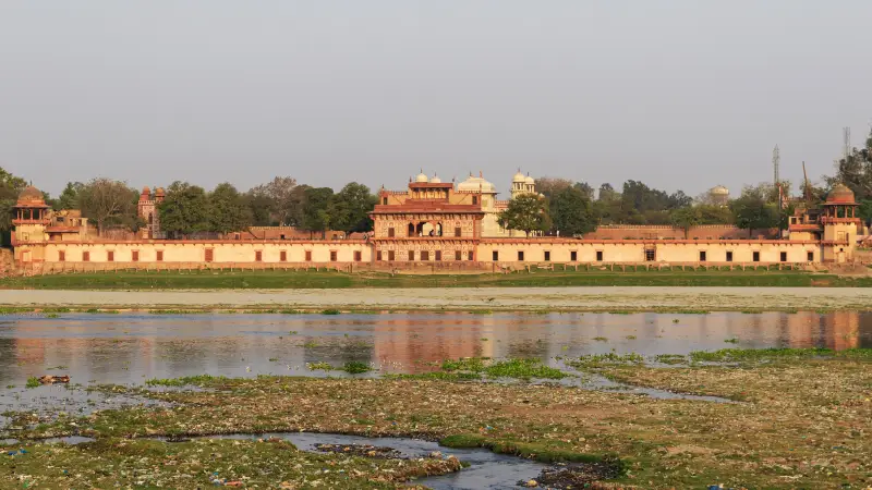 Itimad-Ud-Daulah 2 Panoramic view of the riverside entrance to the Tomb of I'timād-ud-Daulah in Agra, India, taken from across the Yamuna River. The Mughal-era mausoleum, often referred to as the "Baby Taj," is seen with its ornate gateway, symmetrical architecture, and domed pavilions. The riverbank in the foreground is strewn with water plants and visible litter, reflecting environmental concerns around the heritage site.