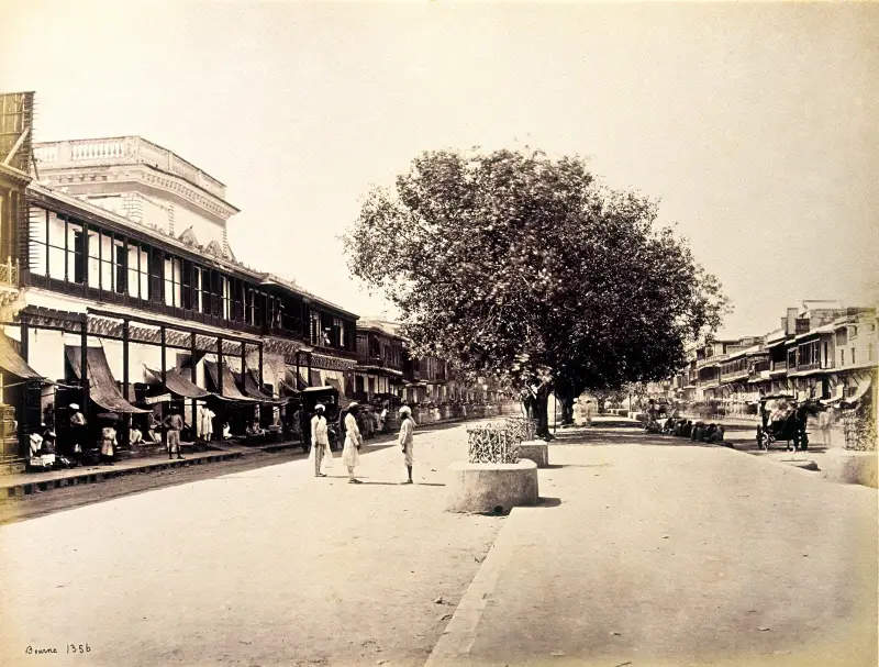 A historic black-and-white photograph of Chandni Chowk, Delhi, taken between 1863 and 1867. The wide street is lined with colonial-style buildings and traditional shops with awnings. A large tree stands prominently in the center, offering shade. A few men in traditional attire walk leisurely on the broad avenue, while horse-drawn carriages are visible in the background, capturing a serene and orderly snapshot of 19th-century urban life in India.