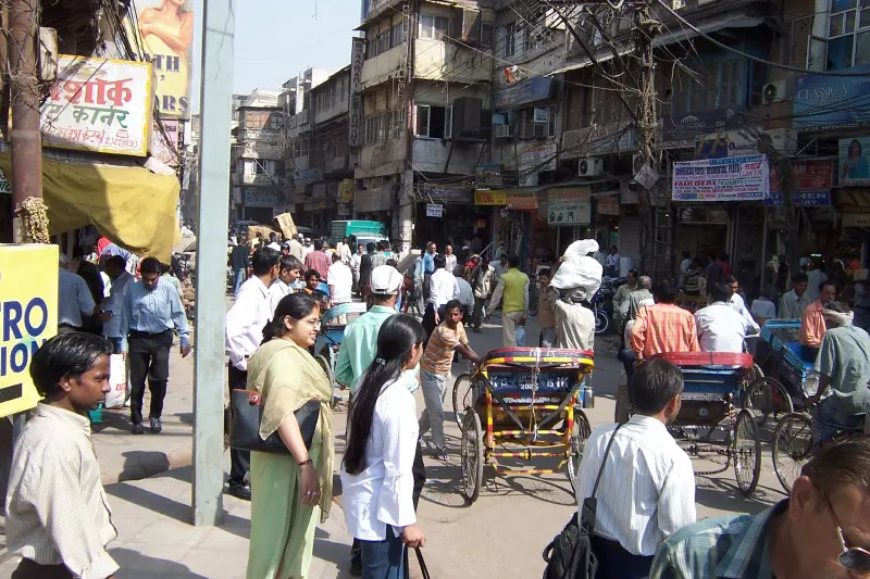 A bustling street scene from Chawri Bazar in Old Delhi, India, filled with pedestrians, cycle rickshaws, and roadside vendors. The narrow street is flanked by aging multi-story buildings packed with signboards and electrical wiring. People from various walks of life navigate the crowded area under the bright sun, highlighting the vibrant, chaotic, and energetic atmosphere typical of this historic marketplace.