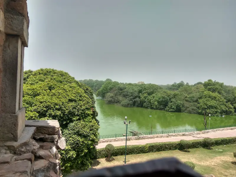 Hauz Khas 2 View of the greenish Hauz Khas Lake in Delhi, surrounded by dense trees and greenery, seen from a stone structure with a portion of a window and wall in the foreground.