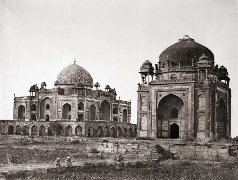Humayun’s Tomb 1 Black and white photograph from 1858 showing Humayun's Tomb in Delhi, India, with the smaller Barber’s Tomb prominently in the foreground. The Mughal architecture features large domes, arched entrances, and intricate stonework. The scene includes an open grassy area with a few seated figures near the steps, highlighting the scale and grandeur of the structures.