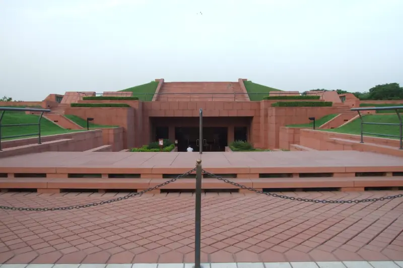 "Exterior view of the Lotus Temple Information Centre in Delhi, India, featuring a red sandstone structure with landscaped green roofs and steps leading to the entrance."
