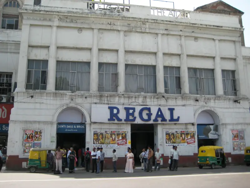 Connaught Place 2 Front view of the historic Regal Cinema building in Connaught Place, New Delhi, with people standing outside and auto rickshaws parked in front. The building has a faded white facade with large columns and movie posters on display.