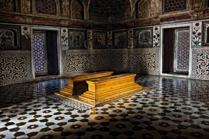 Itimad-Ud-Daulah 1 Interior of the Tomb of I'timād-ud-Daulah in Agra, India, featuring two intricately carved yellow marble cenotaphs set on a polished geometric-patterned marble floor. The walls are adorned with detailed pietra dura floral inlays and lattice windows, showcasing the exquisite craftsmanship of Mughal architecture.