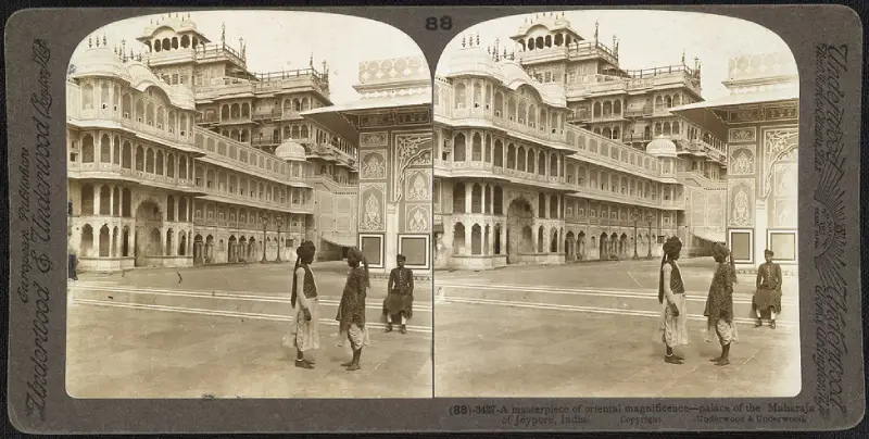 City Palace 1 A historical stereoscopic image showing the grand façade of the City Palace in Jaipur, India, labeled as the palace of the Maharaja of Jeypore. The palace displays intricate Rajput and Mughal architectural styles with multiple balconies, arched windows, and detailed fresco work. In the foreground, traditionally dressed men walk across the spacious courtyard, enhancing the regal ambiance of the scene from the early 20th century.