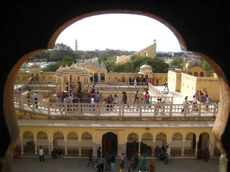 View through an arched window of the inner courtyard of Hawa Mahal in Jaipur, India, showing visitors walking along the ornate upper gallery with domed pavilions, traditional Rajasthani architecture, and cityscape in the background.