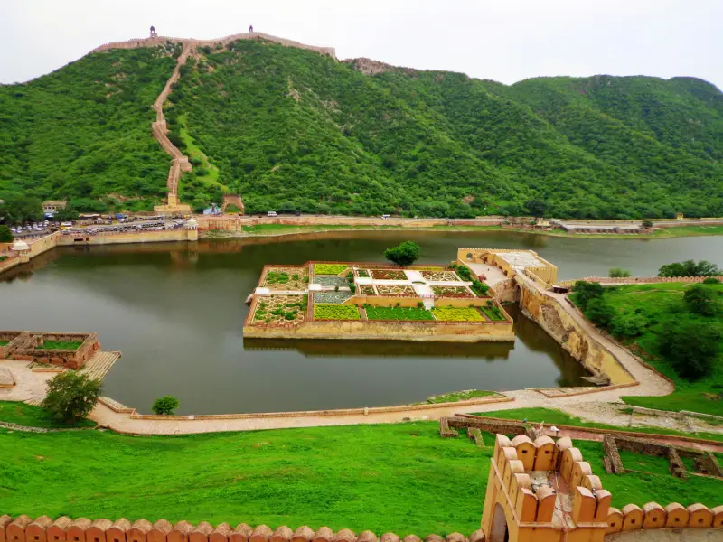 Amer Fort 2 Scenic view of Maota Lake located at the base of Amber Fort in Jaipur, Rajasthan, featuring a symmetrical island garden known as Kesar Kyari Bagh in the center of the lake. The garden is geometrically designed with lush greenery and flowerbeds, accessible via a narrow stone bridge. Surrounding the lake are fortified walls and a steep stone stairway leading up the lush green Aravalli hills toward watchtowers atop the ridgeline, resembling a miniature Great Wall. The landscape showcases the fusion of natural beauty and Rajput architecture.