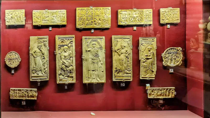 Albert Hall Museum 1 Display case featuring intricately carved ivory or stone reliefs from the Medieval European collection at the Albert Hall Museum. The artifacts are mounted on a deep red background and numbered for identification. The reliefs depict religious and mythological scenes, including figures in robes, angelic beings, and narrative panels. Central panels show full-length human figures in classical poses, while surrounding items include circular medallions and multi-paneled carvings, reflecting Greco-Roman influence. The arrangement emphasizes the fine detail and storytelling of European medieval craftsmanship.