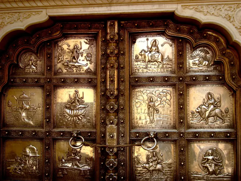 Amer Fort 1 Ornate silver-plated door from Amber Fort, Rajasthan, featuring intricately embossed panels depicting various Hindu deities and mythological scenes. Each rectangular panel contains a distinct religious figure or narrative, framed by detailed floral borders. The door is secured with large circular metal handles and a central locking bar. The craftsmanship showcases traditional Rajasthani artistry with a blend of religious symbolism and decorative elements, set within a grand arched doorway adorned with carved motifs.